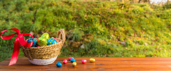 Easter eggs in basket on wooden table banner. Easter season banner with green nature bokeh background. Colorful decorated eggs in wicker basket. Spring season decoration for web banner