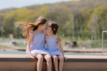 two happy little girls are sitting on a wooden pier on a city lake in summer. Blue sky. Earth Day....