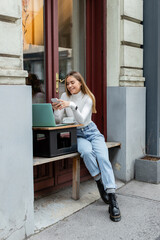 cheerful woman using smartphone near laptop and cup of coffee while sitting in cafe on street in Vienna.