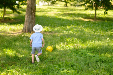 A boy in a headdress, shorts, shirt plays with a yellow ball on the lawn on a sunny summer day.The concept of vacation, vacation, active sports. Copy space.