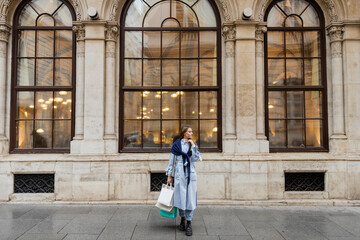 Fototapeta na wymiar young and stylish woman with scarf on top of blue trench coat holding shopping bags near historical building in Vienna.