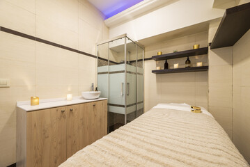 Cabin of a clinic for aesthetic treatments with massage table, shower cabin with glass partitions, wooden sideboard with countertop and white porcelain sink