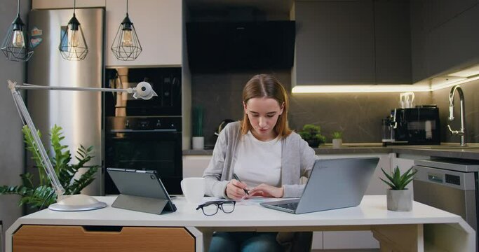 Young female entrepreneur using laptop writing notes in modern at home office desk. Online from home watching web class lesson or listening tutorial. Distance education concept.