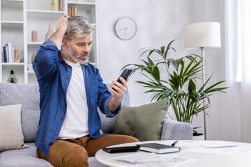 Senior mature adult man sitting on sofa alone at home, using smartphone in living room, reading bad news online from app on phone, upset and nervous.