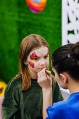 A professional make-up artist, artist paints with a brush on her face with multi-colored paints face painting, drawing, children's makeup for a little girl model. Photography, art concept, lifestyle.