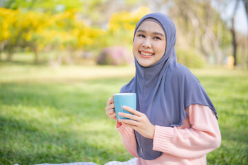 Portrait of Isalamic beautiful woman sitting at garden holding a cup of coffee, teeth smile with hapiness and enjoy her lifestyle relaxing emotion.