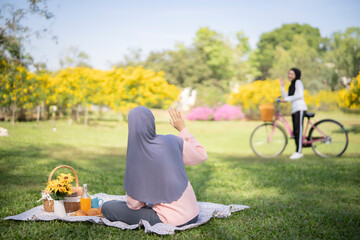 Muslim girl lift her hand to say hi to her friend that riding bicycle far away at park, Islamic...