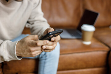 Young man using a smartphone while sitting on sofa at home next to the laptop and a cup of coffee