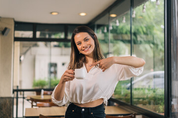 Picture of lovely woman dressed in casual white clothes pointing to cup of coffee to promote it. Positive and good looking young woman takes drinks for advertising content