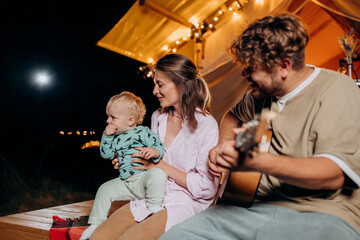 Happy family relaxing and spend time together in glamping on summer evening and playing guitar near cozy bonfire. Luxury camping tent for outdoor recreation and recreation. Lifestyle concept