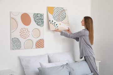 Young woman hanging painting on light wall in bedroom