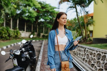 Woman smiling walking in the park outside with laptop freelancer against a backdrop of green palm trees in summer, tropical backdrop, blogger on a trip, work online