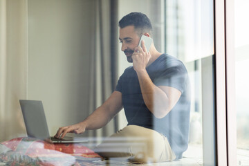 Fototapeta premium Middle aged man talking on cellphone and using laptop, sitting on floor near window at home, free space. Male working on pc
