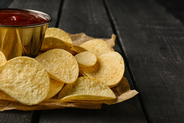 Parchment with delicious potato chips and bowl of ketchup on black wooden background