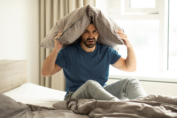 Annoyed caucasian man covering ears with pillow, suffering insomnia, feeling irritated by loud noise, sitting on bed in bedroom, free space