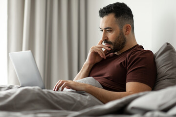 Pensive man using laptop, sitting in bed and thinking while working online at home in bedroom interior, free space. Distance job and technology concept