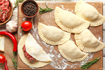 Board with raw meat empanadas and ingredients on light wooden background, closeup