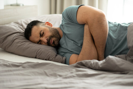 Unhappy Sick Man Lying In Bed And Touching His Belly, Holding Hands On Stomach, Suffering From Pain In The Morning, Home Interior, Copy Space