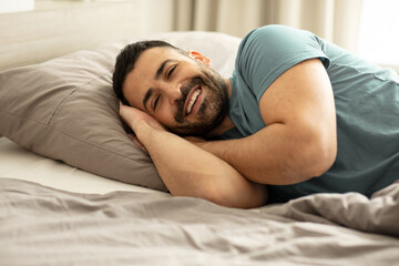 Portrait of happy well-slept man lying in bed, looking and smiling at camera, resting in bedroom in the morning, free copy space