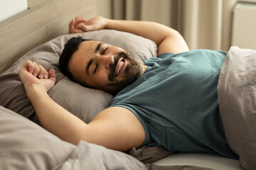 Happy relaxed man lying in comfy bed, enjoying good morning in bedroom, stretching his body after...