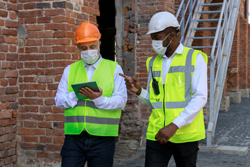 Multicultural coworkers in helmets and face masks discussing construction project details while walking outdoors along unfinished building. Adult caucasian man fixating information on digital tablet.