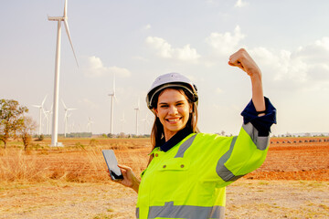 Caucasian female technician holding smartphone working in wind farm field with fists up, happy and...