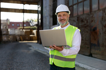 Portrait of the man builder in helmet and vest using wireless laptop for improving blueprints while...