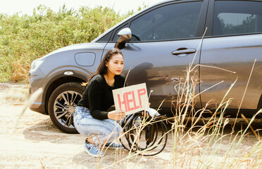 Asian woman sitting next to a car holding cable an electric plug an sign asking for help car...