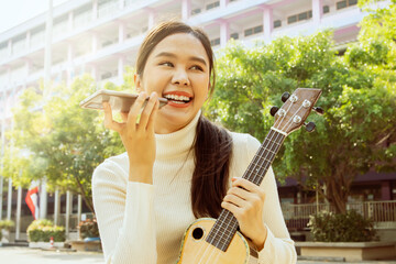 Beautiful Asian teacher sits on ukulele in front of school building preparing to teach students in...