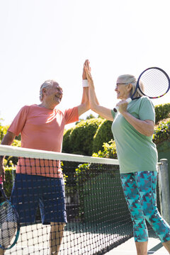 Happy caucasian senior friends high fiving at tennis court on sunny day, copy space