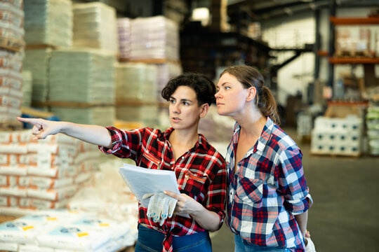 Two women co-workers talking about stock control in warehouse, verifying check list and pointing with finger.