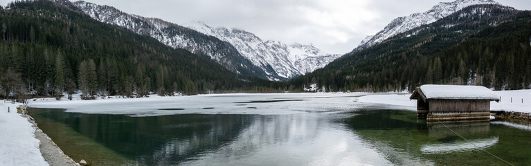 Fototapeta premium Panorama of the Jagersee lake in the Austrian mountains. Frozen water of a lake in the snowy alps with a wooden hut on the water.