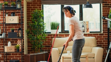 Fototapeta na wymiar Female person listening to music on headset and vacuuming floors, having fun with vacuum cleaner. Young modern woman feeling cheerful while she does spring cleaning household.