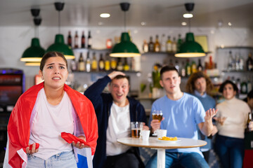 Fototapeta na wymiar Company of upset young adult sports fans holding flag of England and worrying about defeat during game match on TV in pub