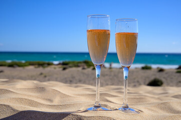 Two glasses of rose champagne or cava sparkling wine served on white sandy tropical beach with dunes and blue ocean, romantic vacation, winter sun on Fuerteventura, Canary, Spain