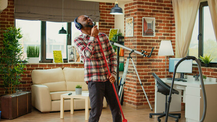 African american person using mop to wash wooden floors, cleaning household and listening to music on headphones. Young smiling man enjoying mopping to clean dirt, household chores.