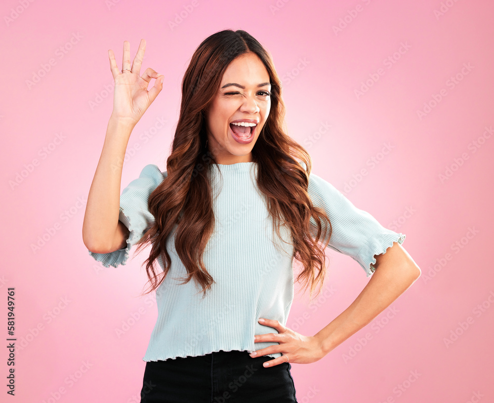 Wall mural Wink, studio and female with a perfect hand gesture, excitement and positive face expression. Happy, smile and young woman model from Colombia with a ok sign language isolated by a pink background.