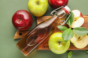 Glass bottle of fresh apple cider vinegar and board with fruits on green background