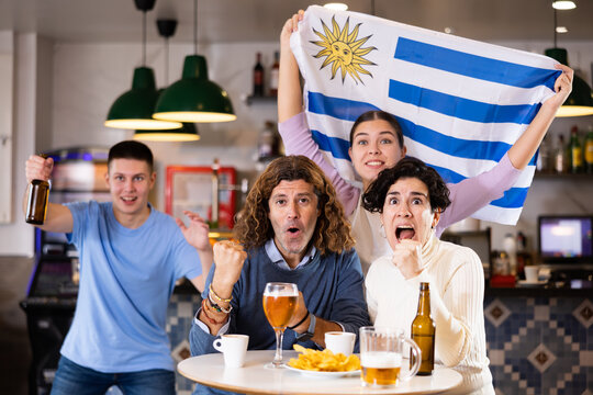 company of emotional young adult sports fans waving the flag of Uruguay and supporting their favorite team with beer in the pub. High quality photo