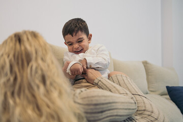 Middle-aged blonde woman lying on the sofa tickling the tummy of her young son with special needs because he is a person with a disability who laughs out loud. - obrazy, fototapety, plakaty