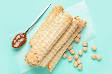 Stack of delicious wafer rolls, spoon with boiled condensed milk and hazelnuts on turquoise background