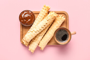 Board with delicious wafer rolls, cup of coffee and boiled condensed milk on pink background