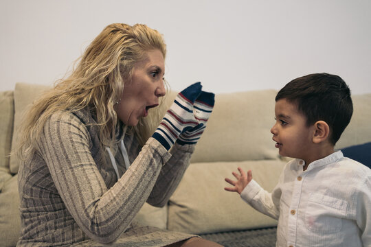 Side image of a middle-aged blonde woman sitting on the couch with socks in her hand playing with her young son who has special needs because he is a person with a disability.