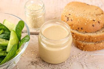 Jar of tasty tahini, salad and bread on light wooden table, closeup