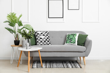 Cozy sofa with cushions and houseplants on tables near white wall