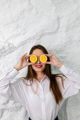 Portrait of a young beautiful brown-eyed girl in a white shirt holding and posing with round slices of lemon. A white wall is in the background. Beauty and Freshness concept.
