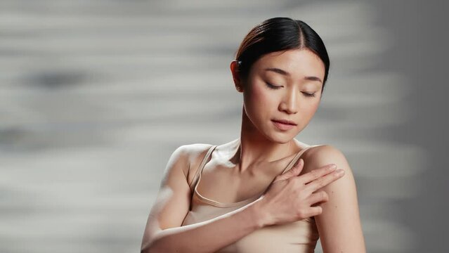 Flawless asian model applying cream on shoulders, promoting body care and positivity in studio. Young woman with confidence creating ad campaign for products in skincare routine.
