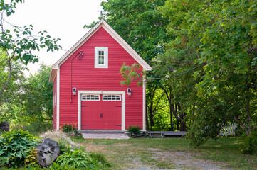 Fototapeta na wymiar A bright red country style red barn or single car garage surrounded by lush green trees and shrubs. The historic building has a small window on the top floor and a garage door on the lower level.
