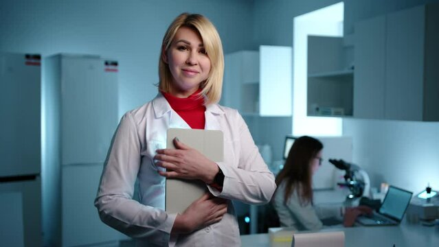 A blode female doctor in a laboratory checking something on the tablet before smiling at the cameraAnother doctor is working on a microscope in the background. High quality 4k footage 