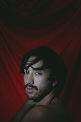 Fototapeta premium Artistic photo of a young latin man looking at camera with bare shoulders and a red cloth background. Vertical photo.